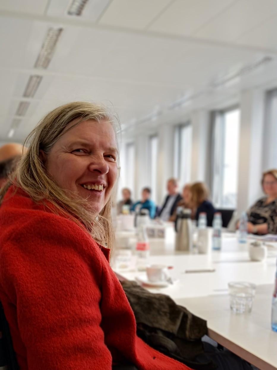 Frau mit blondem langen Haaren und einer roten Jacke sitzt an einem langen Tisch auf dessen Gegenseite weitere Personen zu erkennen sind