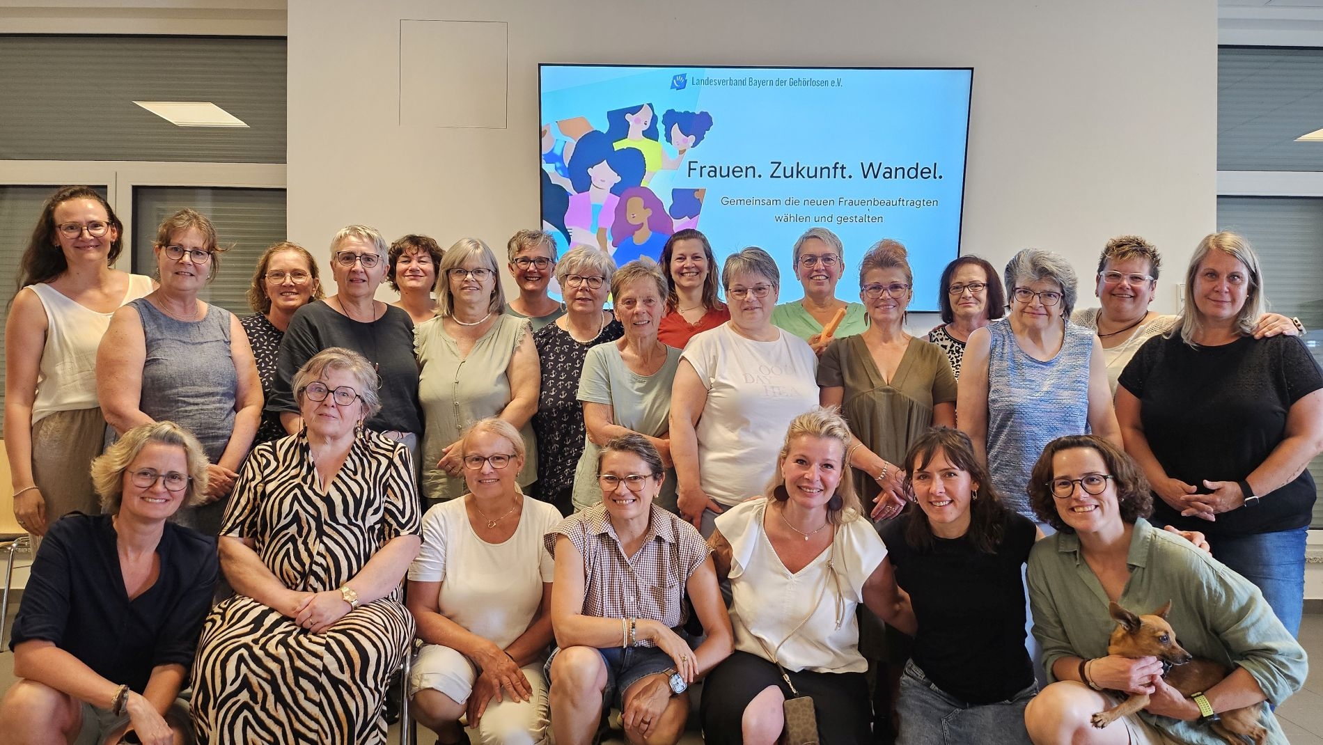 Größer Grupp von Personen vor einer Wand mit dem farbigen Hinweis zu dieser Veranstaltung