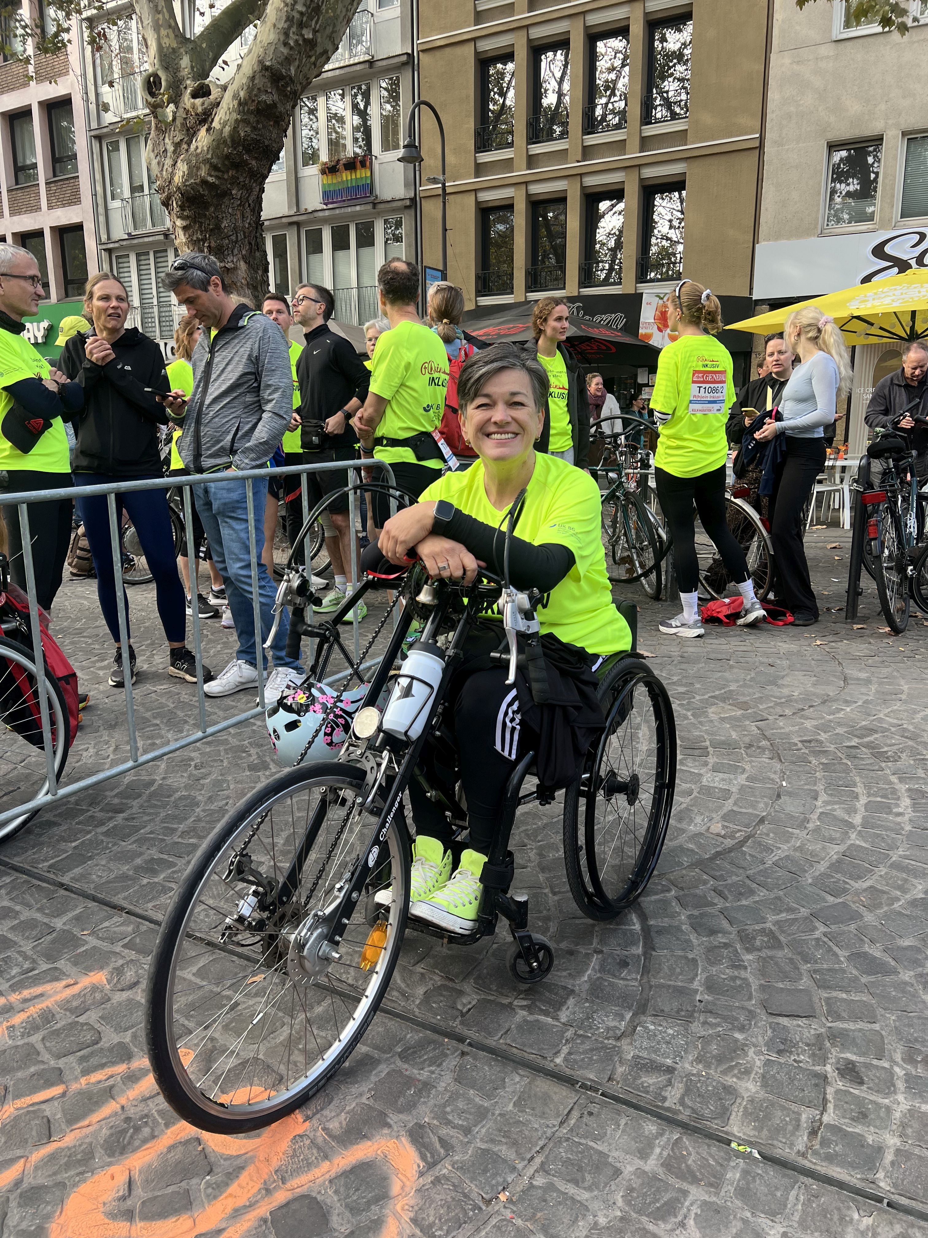 Rollstuhlfahrerin mit Adaptivbike wartet in Wechselzone der Staffel im neongelben „R(h)ein Inklusiv“ T-Shirt