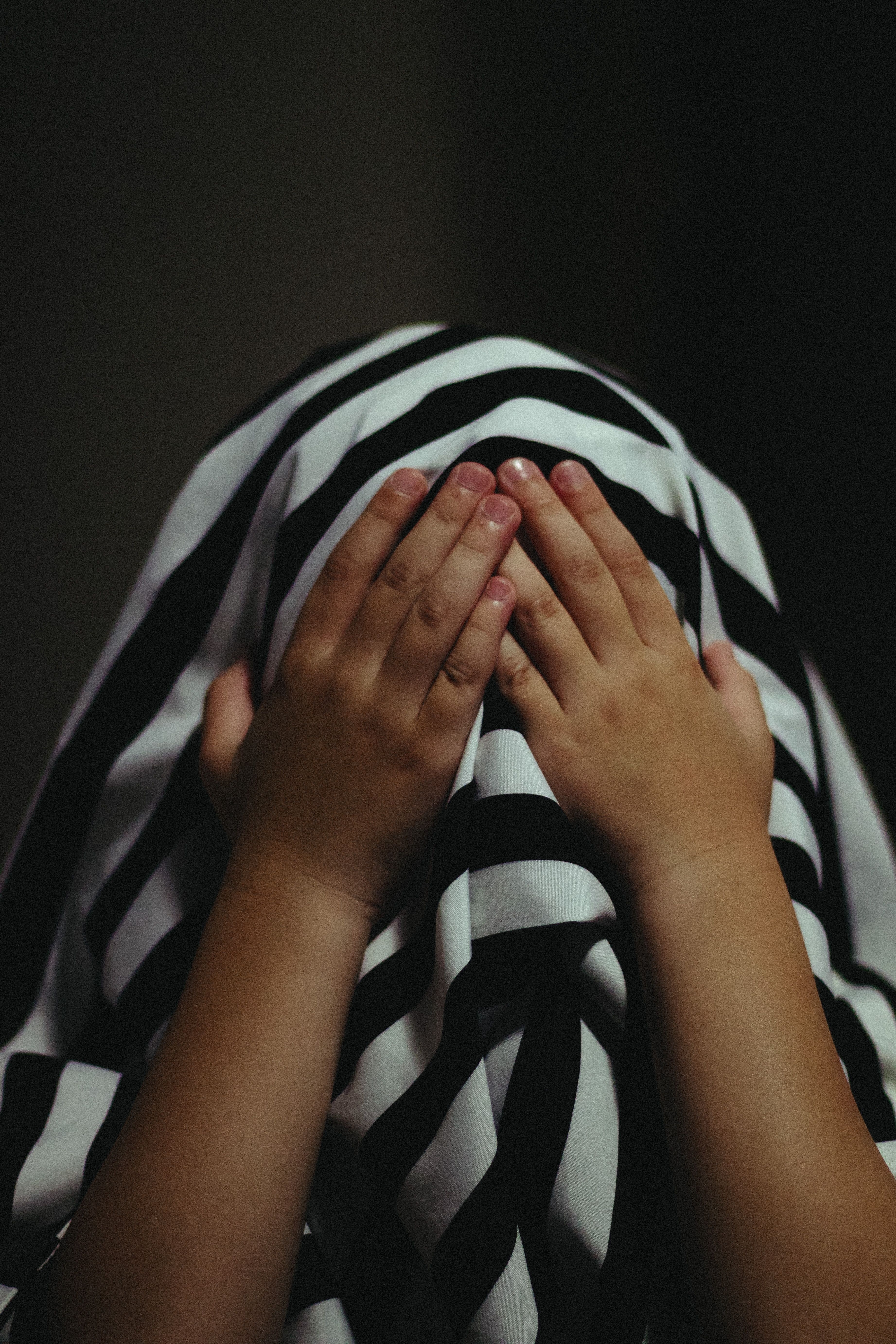 A child covers their face with hands, hidden under a striped cloth against a dark background.