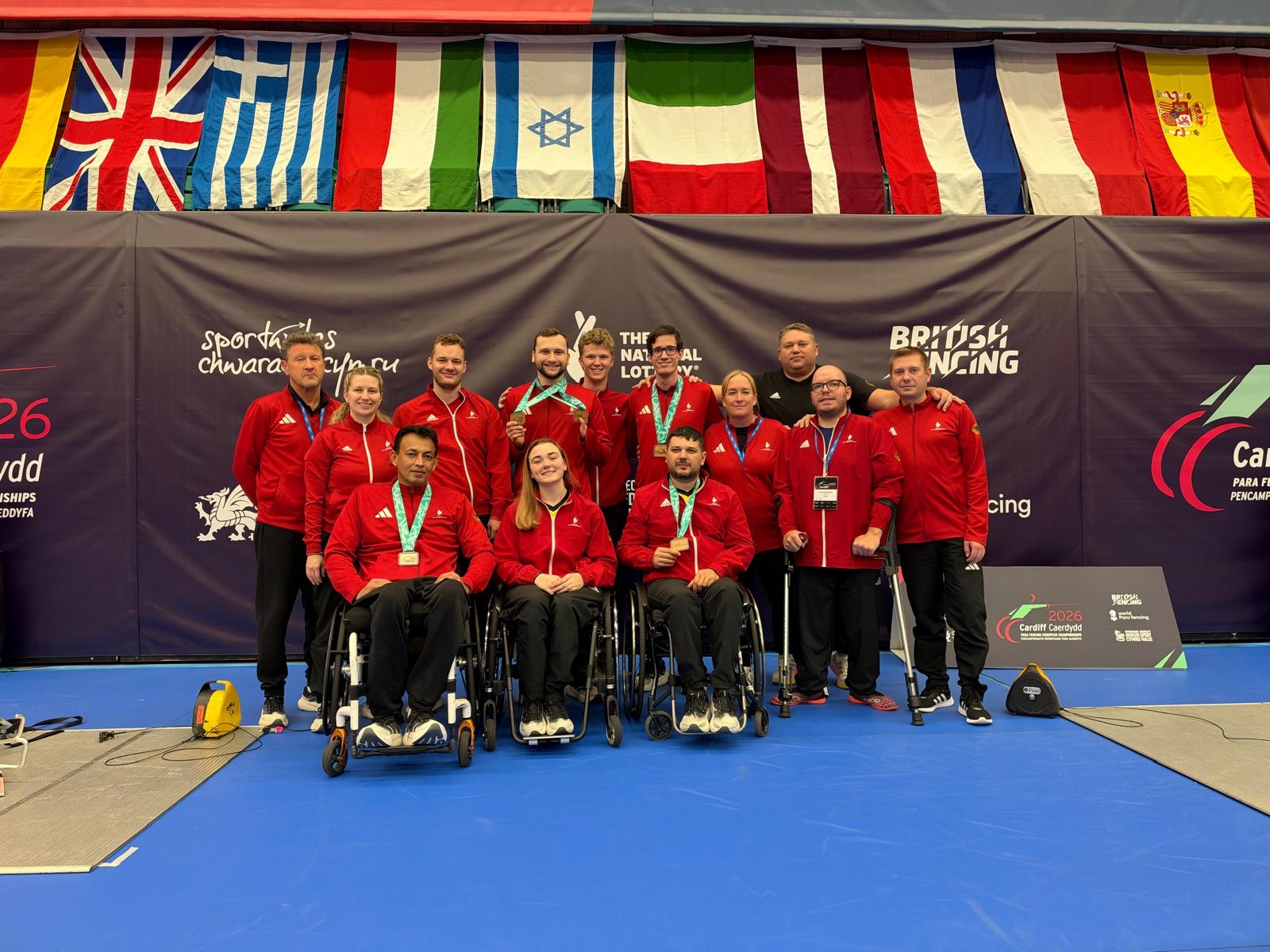 Gruppe von Sportlerinnen und Sportler mit schwarzen Hosen und roten Jacken auf Rollstühlen und in der zweiten reihe stehend