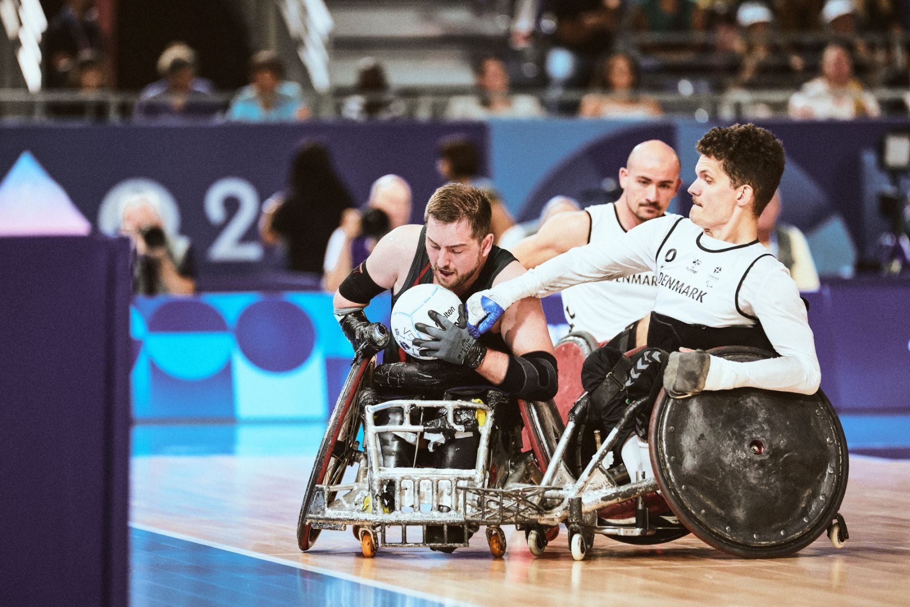 Drei Männer mit Sportrollstühlen, einer im dunklen Trikot, zwei im weißen Trikot beim Spiel