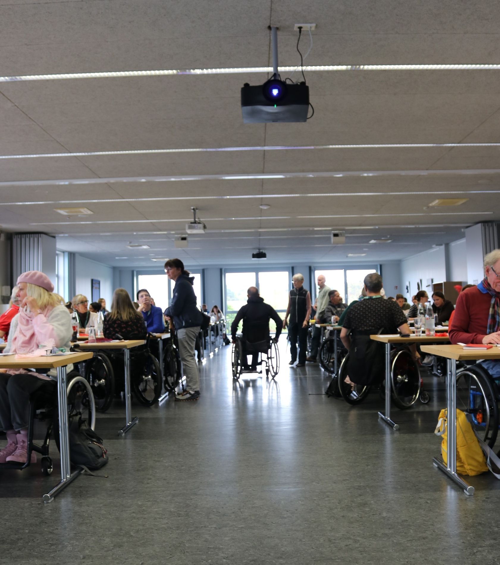 Blick in einen großeg Raum mit Tischen an denen Leute mit Rollstühlen sitzen