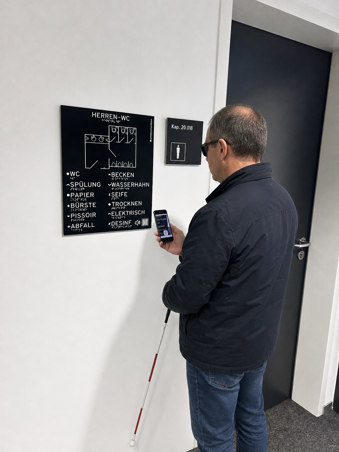 Ein Mann mit Taststock steh vor einem Tastbild an einer Tür und hält ein Smartphone in der Hand