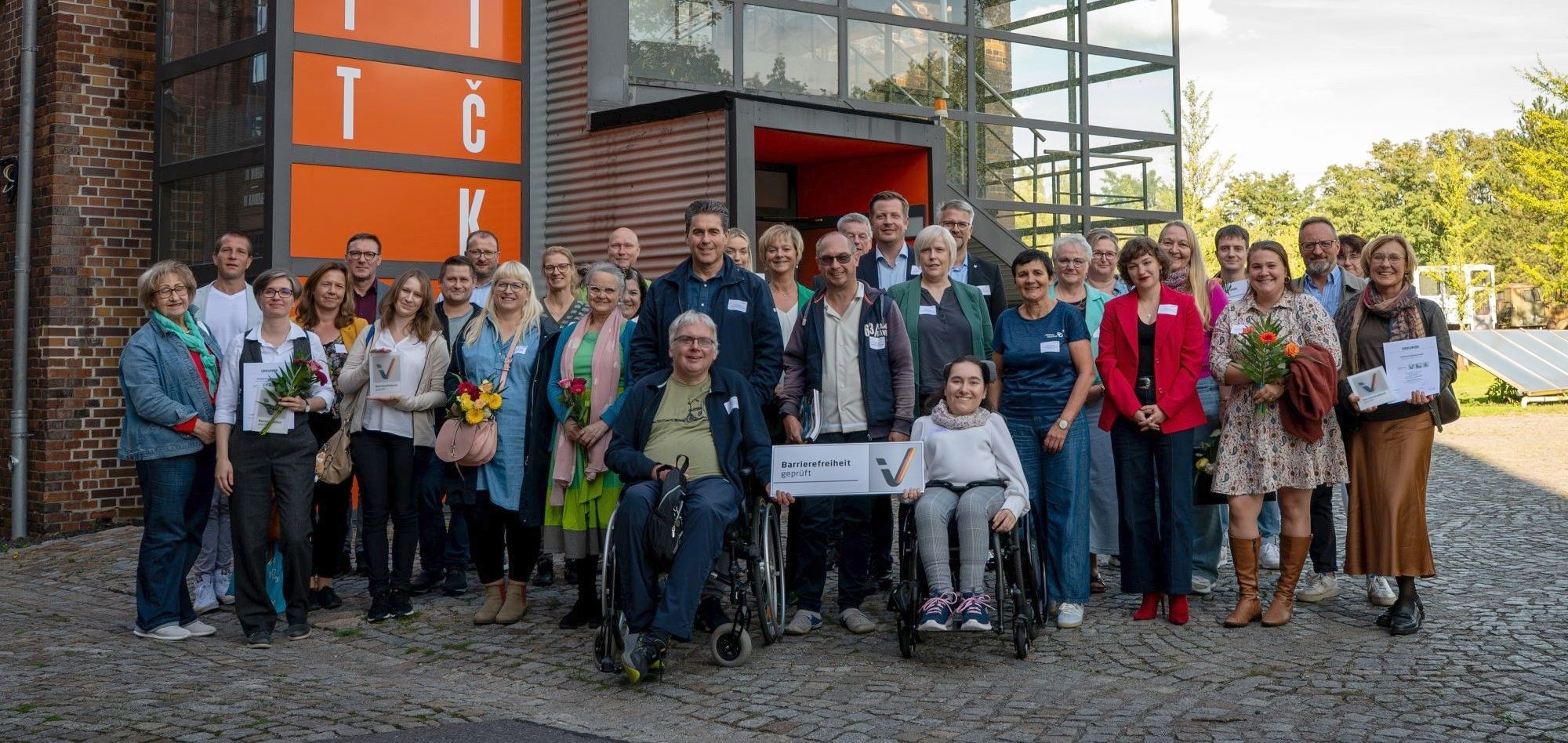 Gruppe von Männern und Frauen, darunter auch zwei Personen mit Rollstuhl vor einer gläseren Eingangshalle