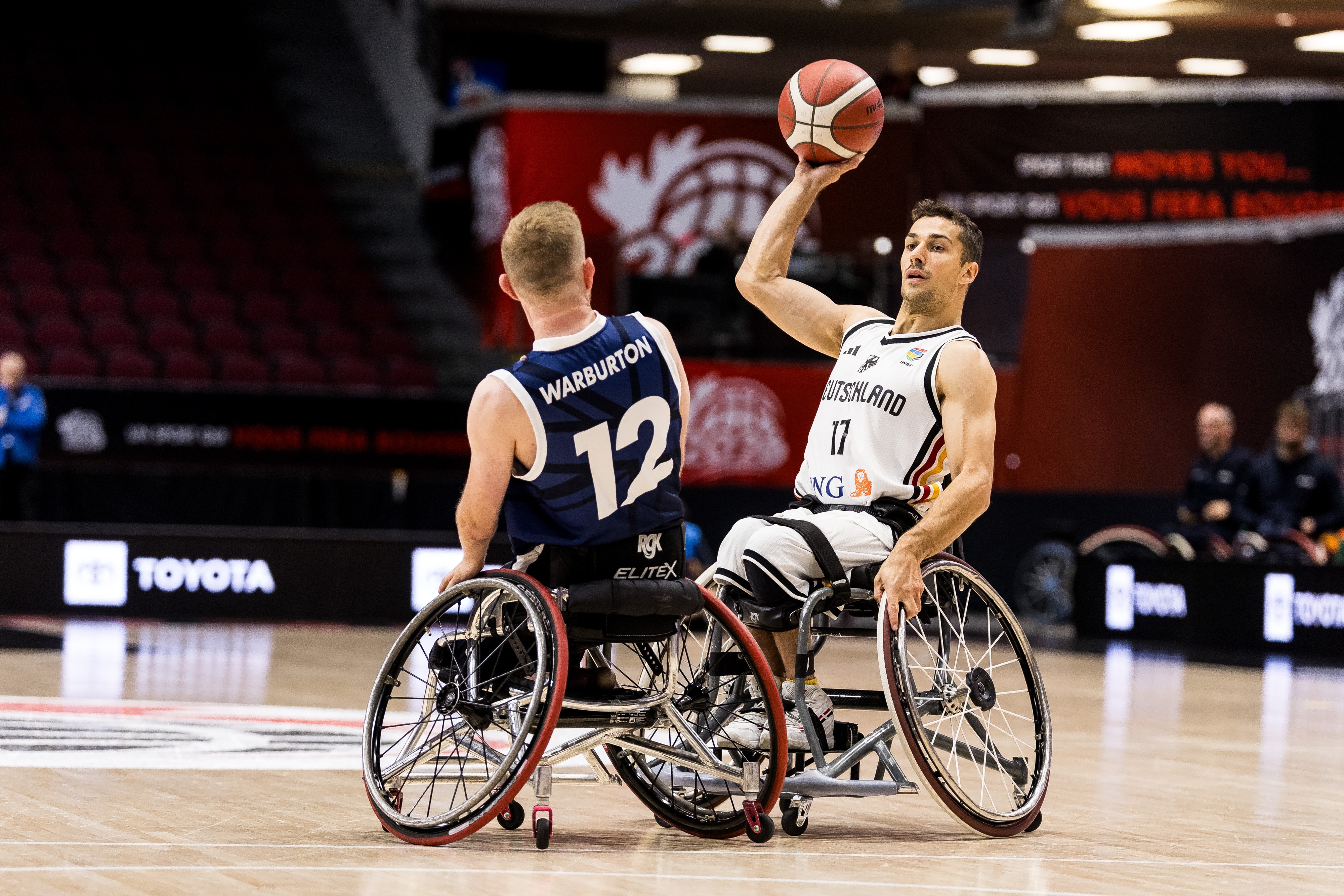 Zwei Rollstuhlbasketballer, davon einer im weißen Trikot mit dem Ball in der Hand, beim Spiel