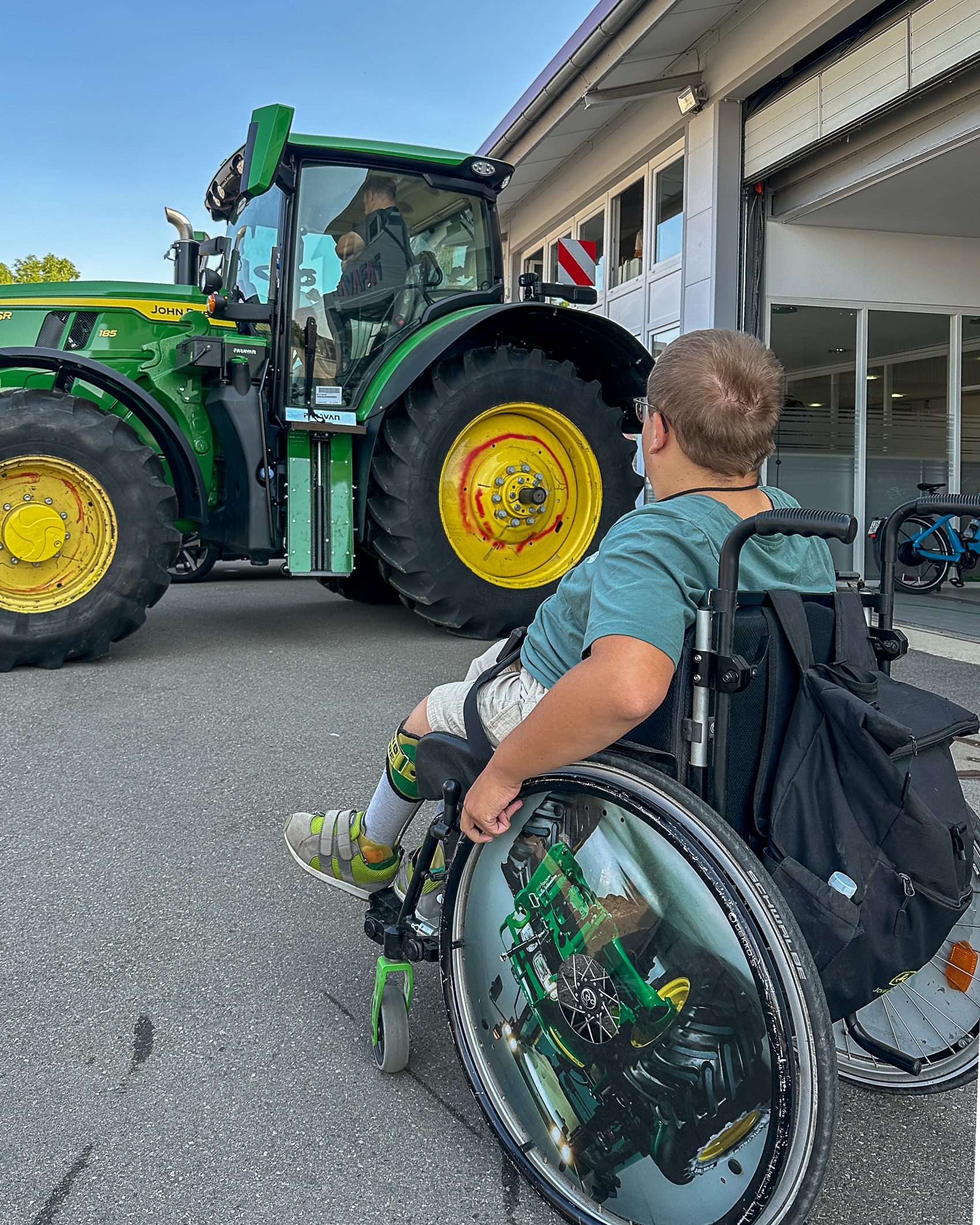 Im Hintergrund ein grüner Traktor mit großen gelben Rädern . Davon ein junger Mann in einem Rollstuhl