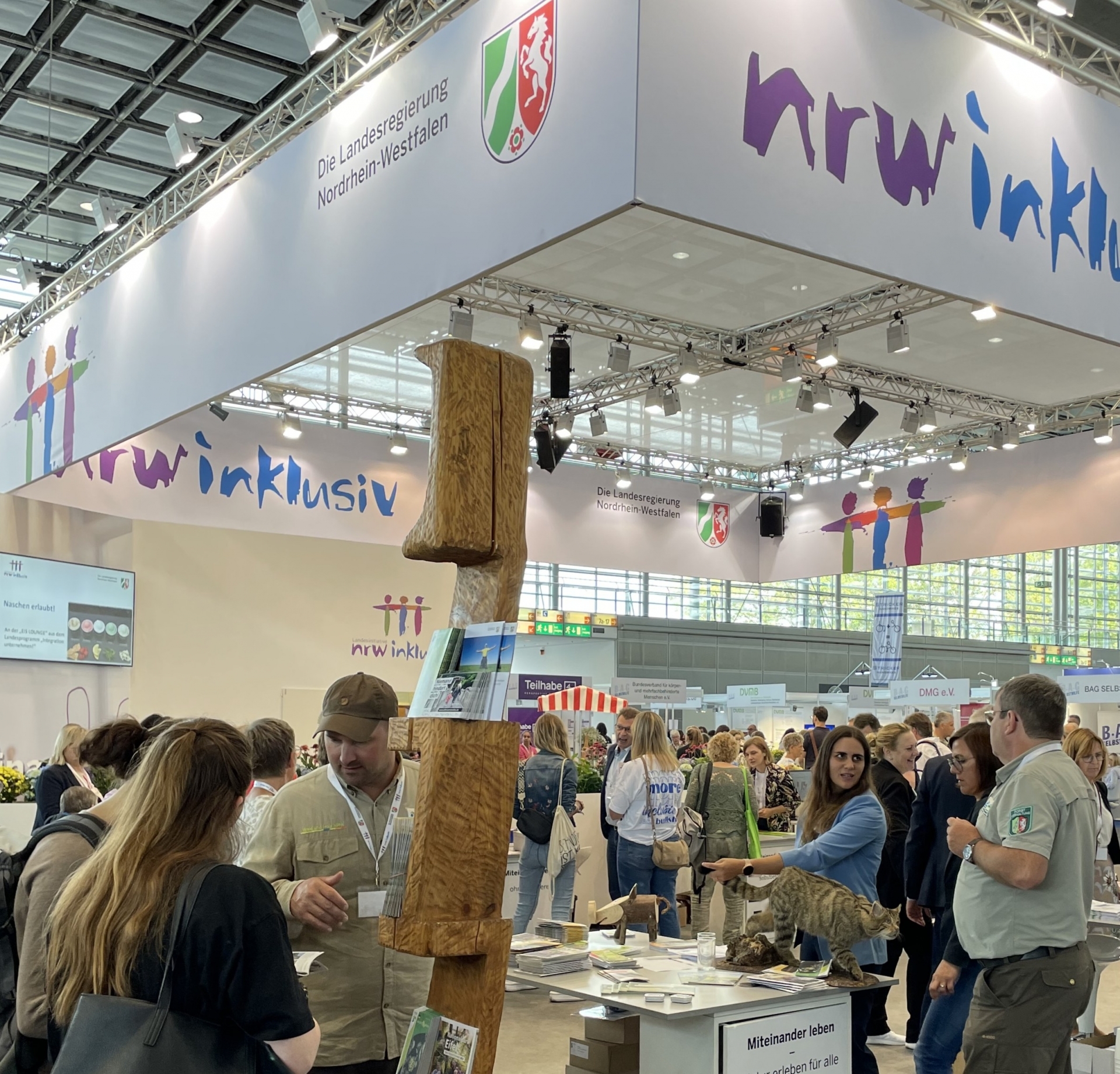 Hell erleuchrtetet quadraatischer Stand einer Messe mit Ständen sowie vielen Besucherinnen und Besuchern