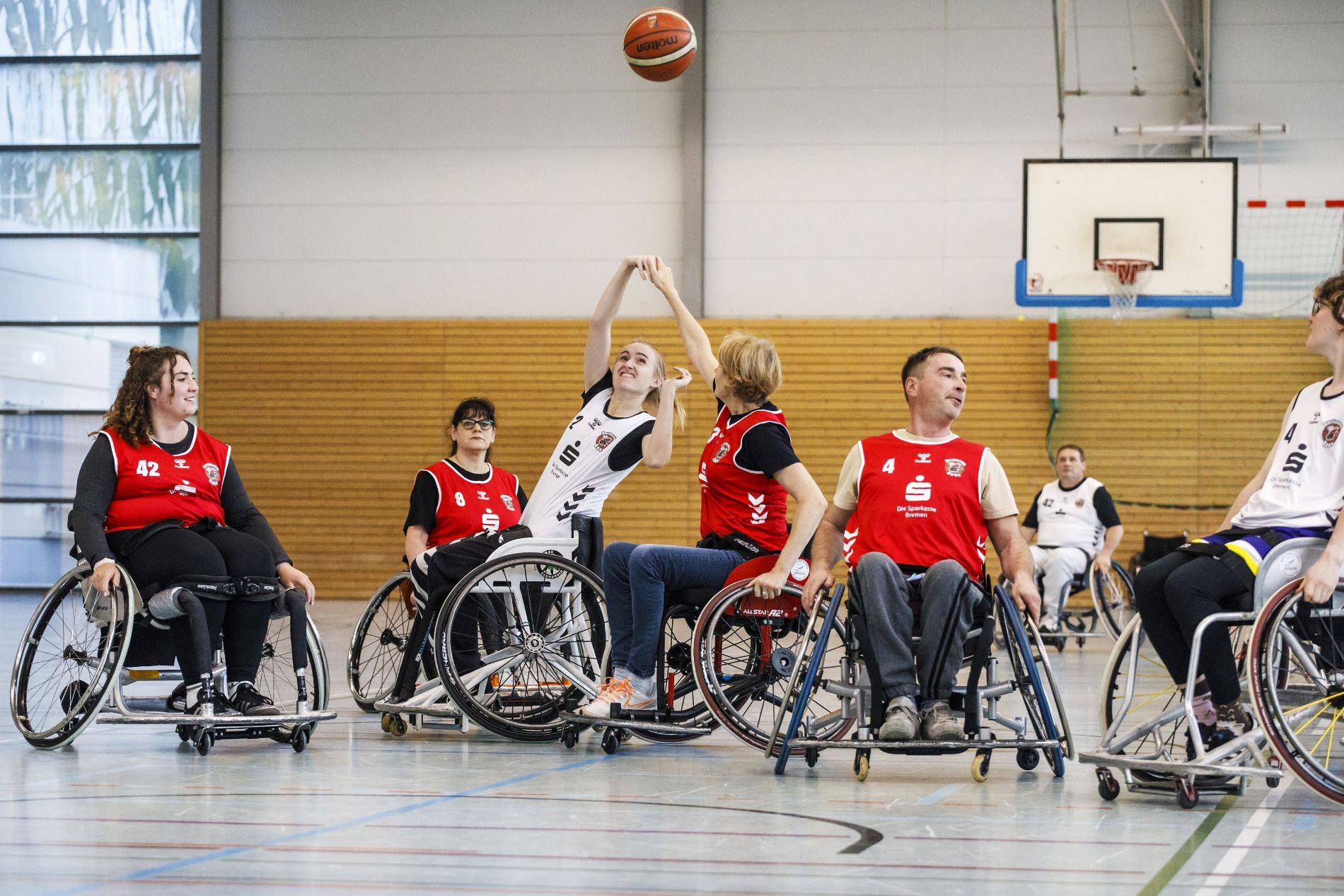 Junge Männer und Frauen in rotem oder weißen Sportdress mit Sportrollstühlen beim Basketballspiel