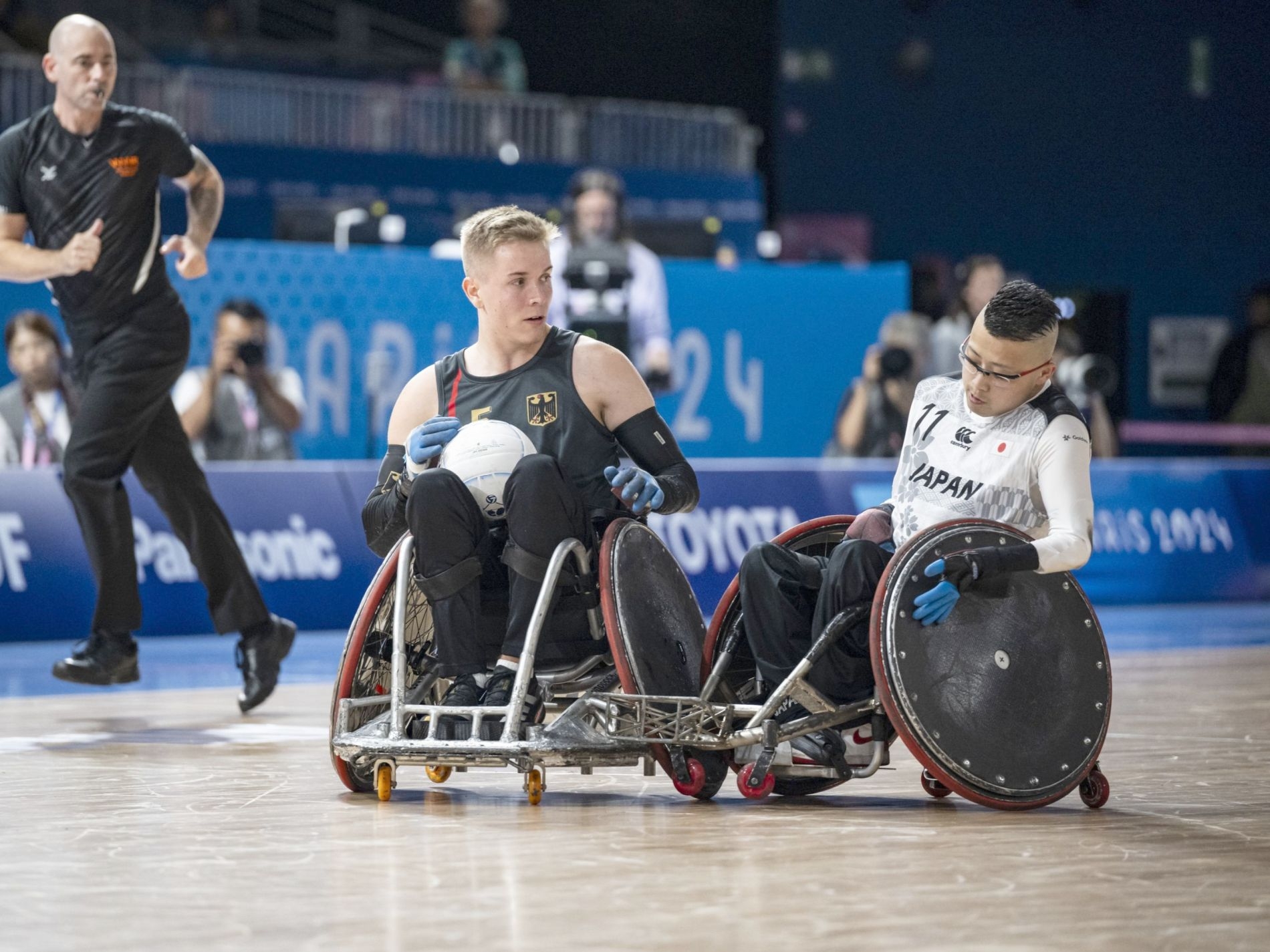 Zwei junge Männer, einer in schwarzem und einer im weißen Sportdress, auf Sportrollstühlen. Einer vo9n beiden hat einen Ball