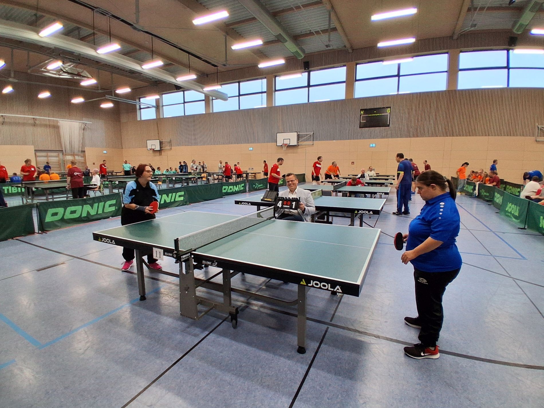 In einer großen Sporthalle stehen mehrere Tischtennisplatten an denen Tischtennis gespielt wird