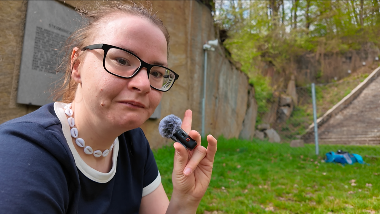 Junge Frau mit Brille sowie glatt nach hinten gekämmten Haar und einem kleinem Mikrofon in der Hand