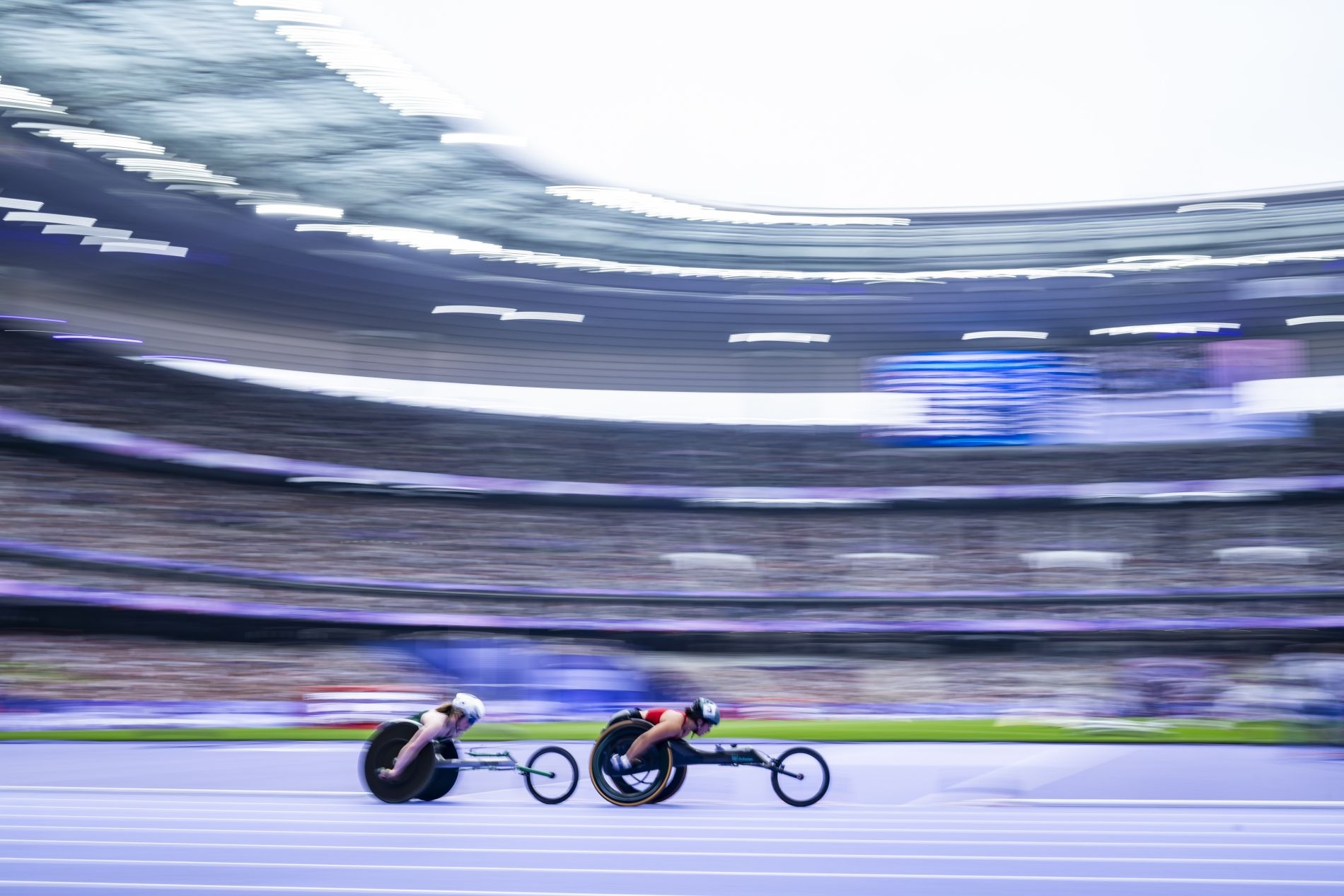 Zwei Sportler auf Rollstuhlrennrädern fahren mit hoher Geschwindigkeit in einem großen Stadium. Die hohe Geschwindigkeit wird durch eine verwischte Stadiumsilhouette anschaulich.