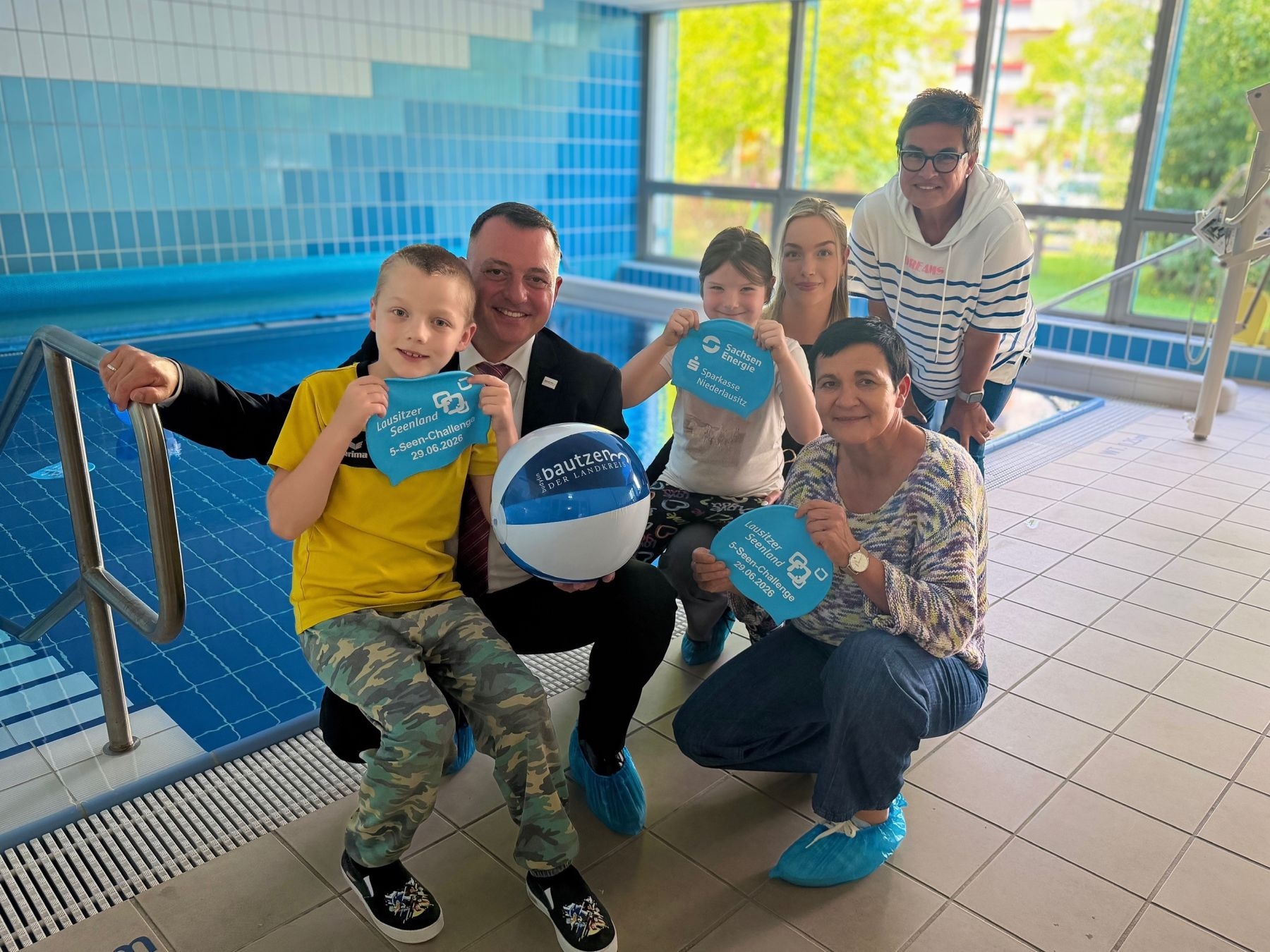 Vier Erwachsene und zwei Kinder an einem Schwimmbecken mit blauen Badekappen in den Händen