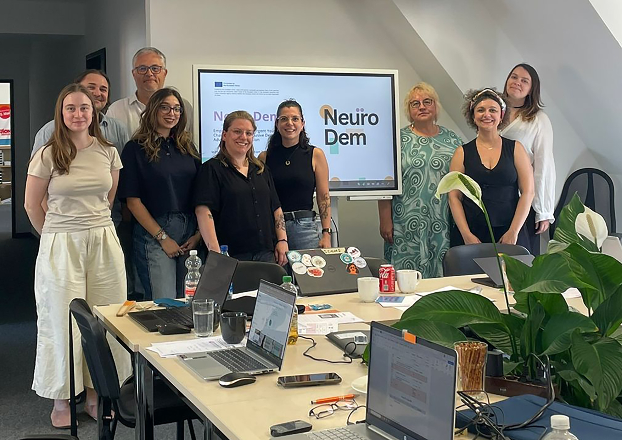 Fotografie einer Gruppe aus acht Personen bei einem Arbeitstreffen in einem Büro mit Bildschirmpräsentation „NeuroDem“. Mehrere Laptops, Notizen und Getränke auf Konferenztisch sowie Pflanzen und Präsentationsmaterial im Raum vermitteln wissenschaftlichen Projekt- oder Teamkontext.
