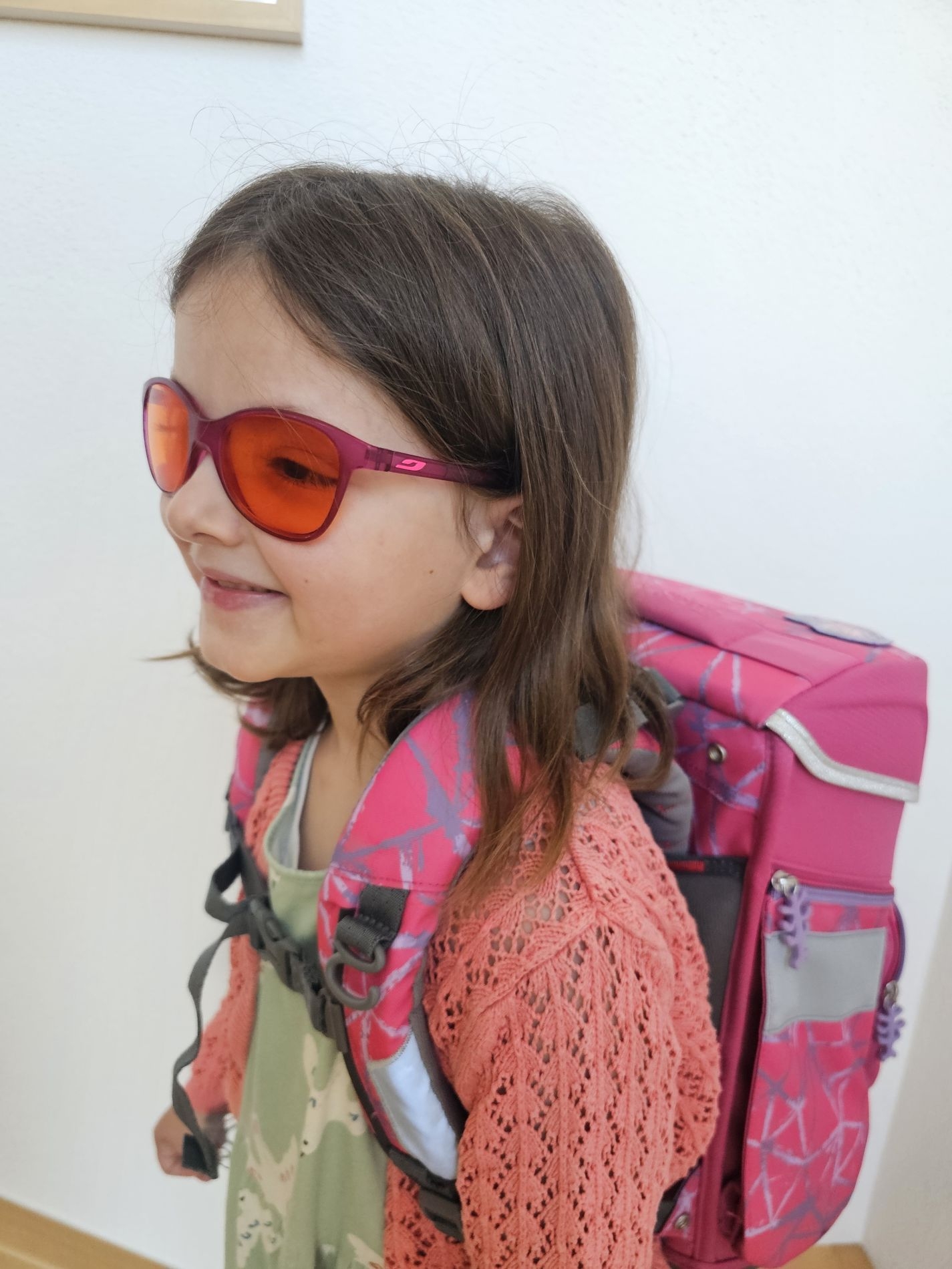 Kleines Mädchen mit dunklem Haar und dunkler Brille mit einem pinken Schulranzen