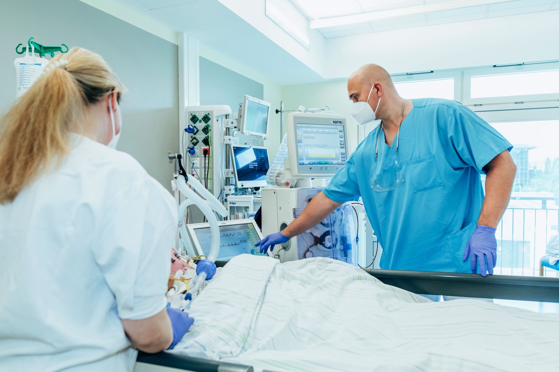 Frau mit blondem Haar in weißem Kittel steht an einem Krankenbett, auf dem ein Patient liegt. An der anderen Seite des Bettes ein Mann in blauem Kittel und mit gesichtsmaske