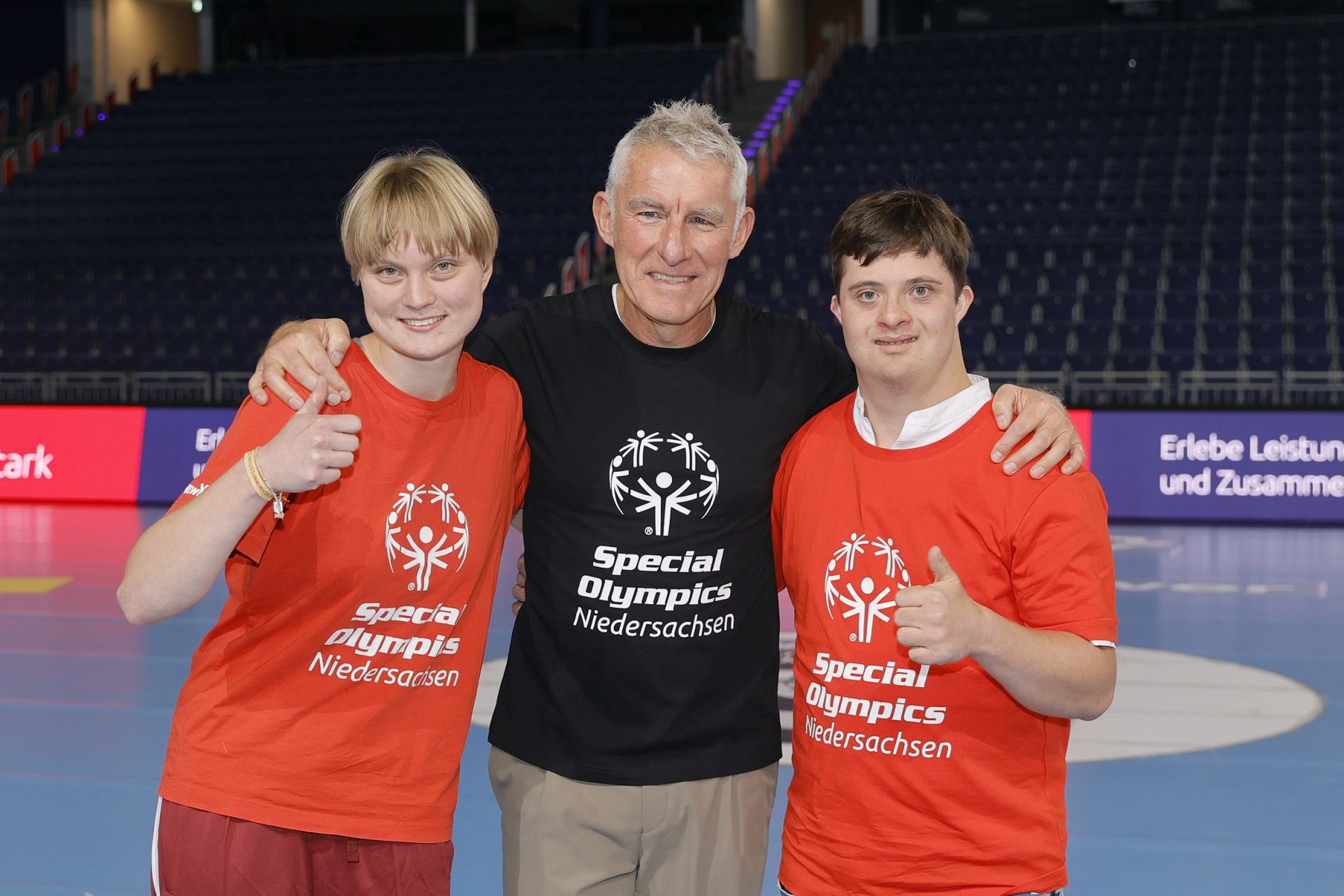 Eine junge Frau mit kurzem Haar und ein junger Mann mit roten Shirt der Special OLympics sowie in der Mitte ein Mann mit schwarzem Shirt der Special Olympics