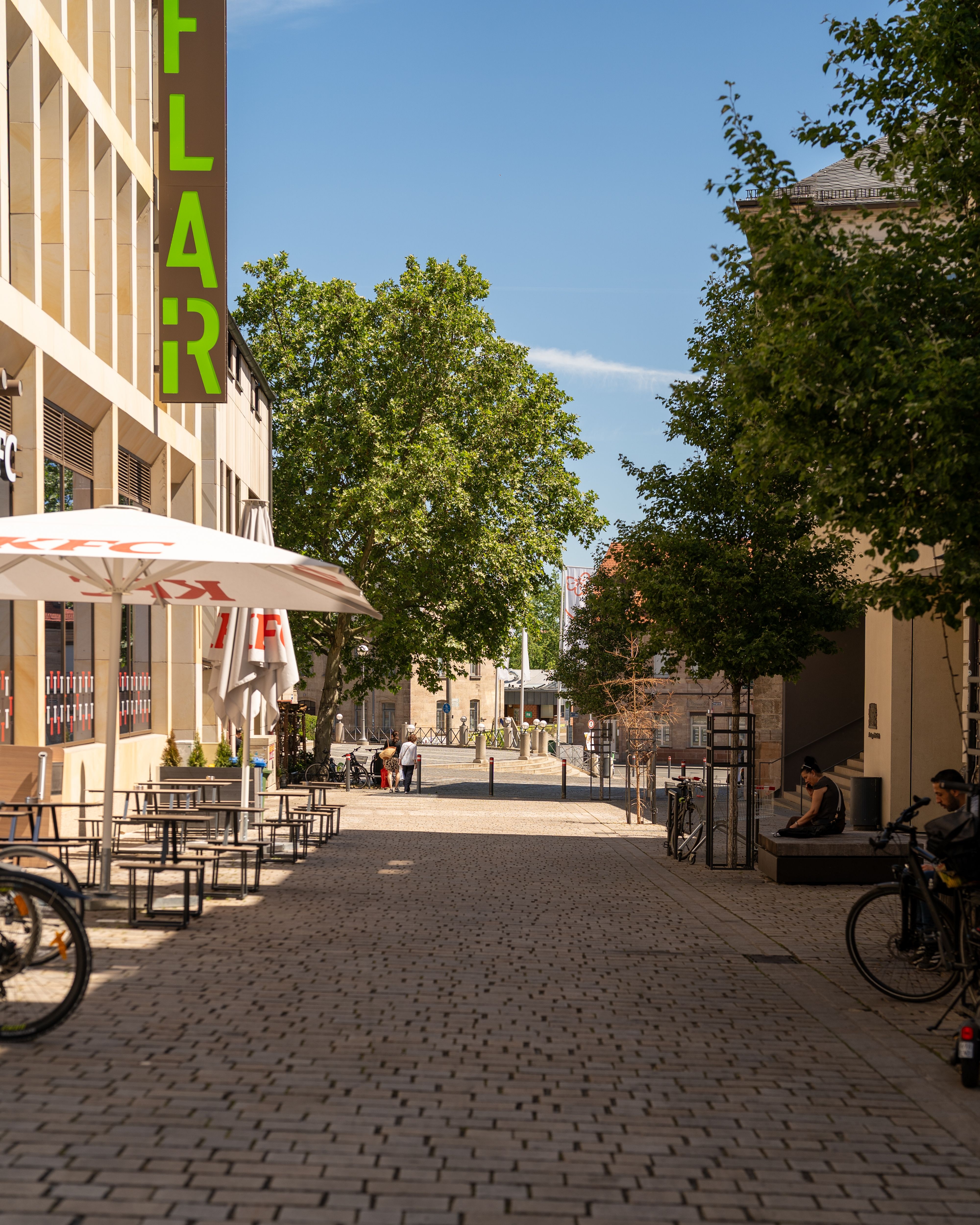 Charming urban street with outdoor seating and lush greenery under a clear sky.
